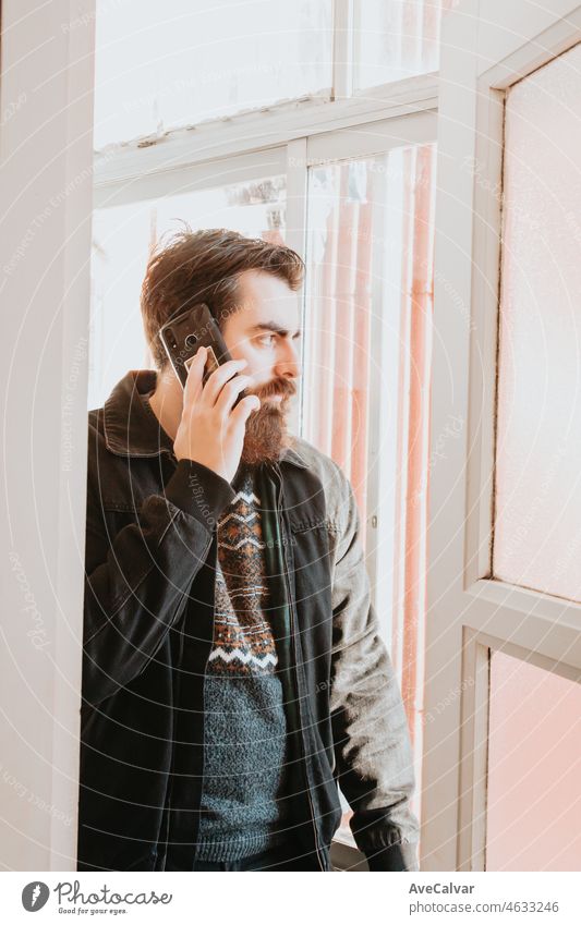 Hübscher junger bärtiger Mann, der telefoniert und sein Telefon am Fenster seines Hauses überprüft. Schaut aus dem Fenster, während er mit seinem Handy telefoniert. Langer Bart und Hipster-Stil