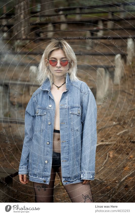 Ein wunderschönes blondes Mädchen steht vor einer Kamera. Mit ihrer blauen Jeansjacke, der roten Sonnenbrille, dem Tattoo und dem Piercing. Etwas Rauch umgibt sie schön.