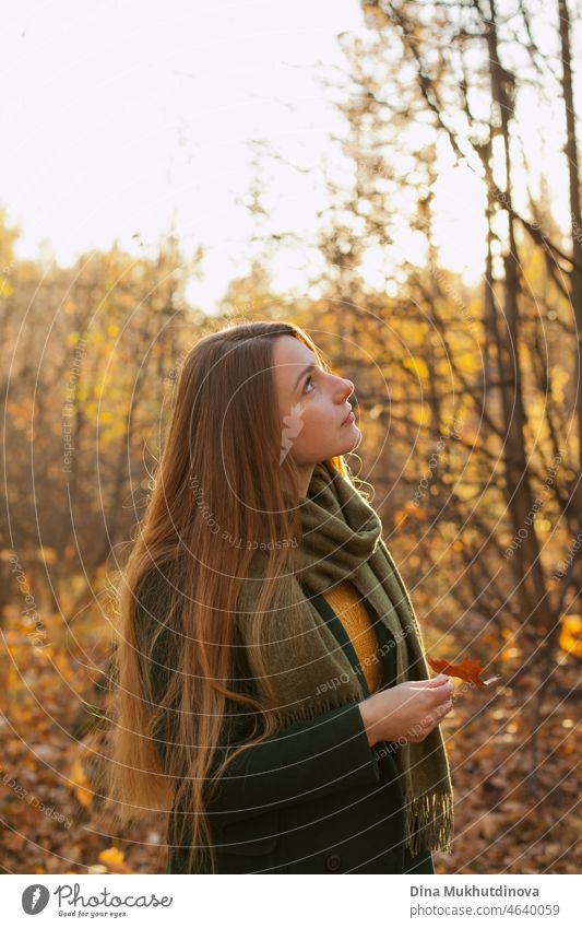 Schöne Frau mit langen Haaren in grünem Mantel steht im Herbst Park hält einen Herbst Ahornblatt. Stil, Mode und Kleidung. Tief in Gedanken im Herbst Wald in der Natur. Authentischer Lebensstil.