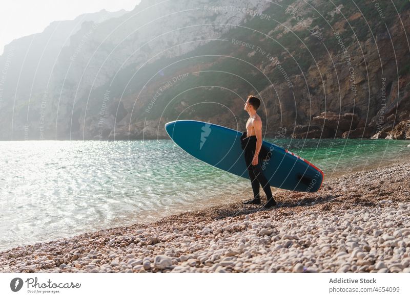 Mann mit Paddelbrett am steinigen Meeresufer an einem sonnigen Morgen Surfer Surfen Surfbrett MEER Wasser Zusatzplatine Küste Strand Hobby Brandung Aktivität