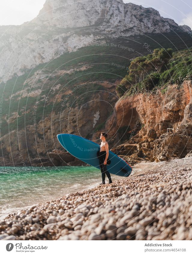 Mann mit Paddelbrett am steinigen Meeresufer an einem sonnigen Morgen Surfer Surfen Surfbrett MEER Wasser Zusatzplatine Küste Strand Hobby Brandung Aktivität