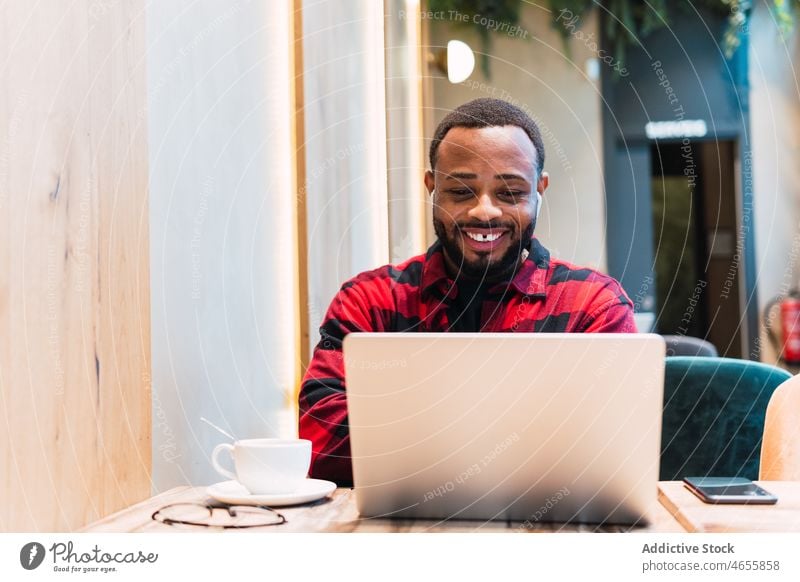 Lächelnder schwarzer Mann arbeitet an einem Laptop in einem Café Kopfhörer benutzend online Internet Browsen freiberuflich Arbeit männlich Afroamerikaner