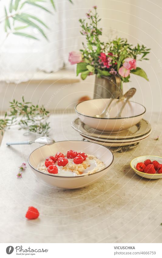 Weiße Schüssel mit veganem Porridge mit Himbeeren am weißen Betonküchentisch Schalen & Schüsseln Veganer Haferbrei Küche Tisch Geschirr Blume Haufen Fenster