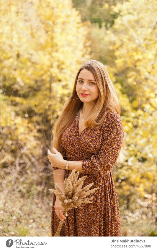 Schöne Frau im Herbst Park trägt ein braunes Kleid, hält einen trockenen Weizen Bouquet. Junge Millennial-Frau mit langen Haaren in stilvollen Herbst-Outfit, lächelnd und Blick in die Kamera.