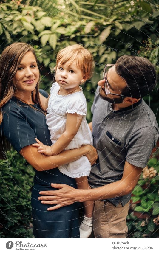 Kleines Mädchen in den Armen ihrer Eltern. Familie genießt im Garten. Hübsche blonde Kinder zwischen ihren jungen Eltern. Liebe und Familie Konzept. Papa Mama