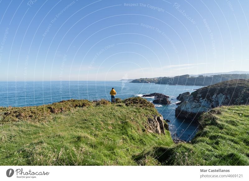 Küstenlandschaft und Mädchen in gelbem Kapuzenmantel, das aufs Meer schaut und spazieren geht. Raum kopieren. Landschaft MEER Himmel Küstenlinie Frau Person