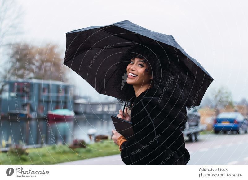 Lächelnde schwarze Frau mit Regenschirm auf der Straße Großstadt Straßenrand urban Vorschein Tablette Porträt afroamerikanische Frau lässig jung Glück positiv