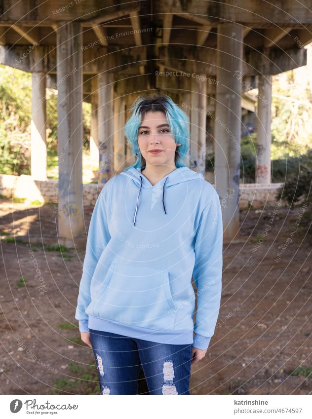 Blauhaariges lächelndes Teenager-Mädchen in hellblauem Oversize-Kapuzenpulli bleibt unter Brücke Lächeln hell-blau Attrappe Übergröße Jeanshose blauhaarig