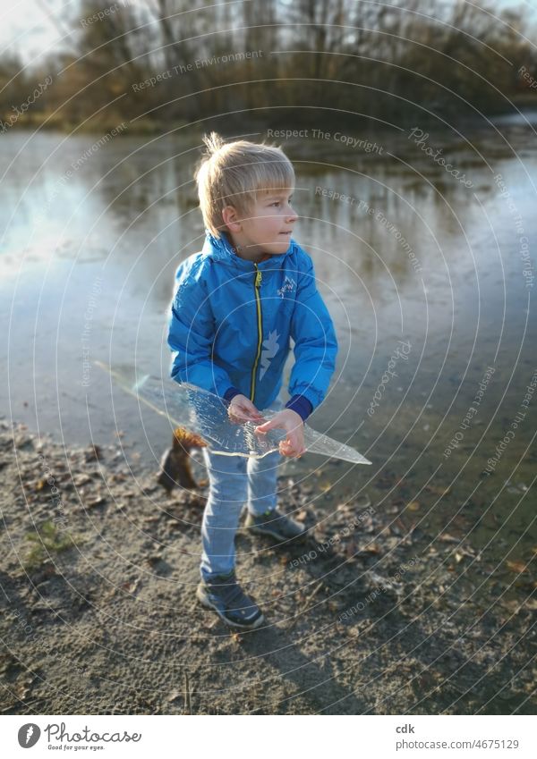 Kindheit | Naturerfahrung | eiskalte Angelegenheit. Junge blond blau blauer Anorak blaue Jeans See Weiher Eisscholle Eisplatte Wasser Sand werfen halten draußen