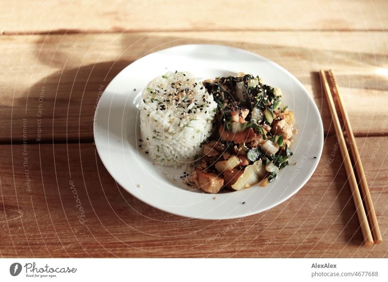 Suppenteller mit einem asiatisch Gericht mit Reis, Huhn, Pak Choi und Sesam auf einem Holztisch mit Essstäbchen Teller schwarzer Sesam gelber Sesam Wokgericht