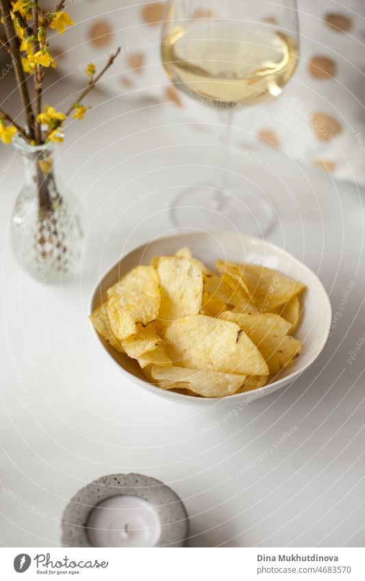 Einzelne Weißweinglas und Kartoffelchips auf weißem Tisch zu Hause. Einzelne Wohnung leben. Home Interieur mit gelber Couch. Entspannender Abend allein zu Hause. Ein-Personen-Haushalte werden weltweit immer häufiger. Lebensstil der Millennials.