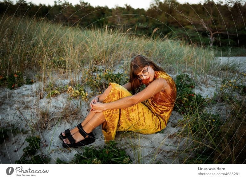 An einem perfekten Sommerabend zur goldenen Stunde macht es sich ein wunderschönes brünettes Mädchen in einem goldenen Outfit ganz allein in den goldenen Dünen gemütlich. Es ist ein majestätischer Sonnenuntergang, der ihr hübsches Gesicht in Gold färbt. Und auch die Goldpreise steigen.