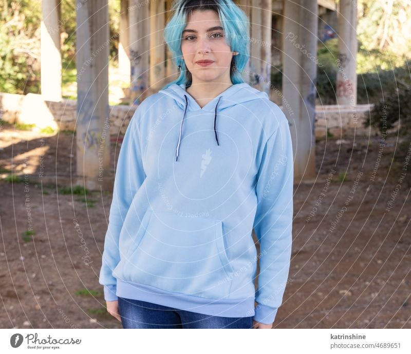 Blauhaariges lächelndes Teenager-Mädchen in hellblauem Oversize-Kapuzenpulli bleibt unter Brücke Lächeln hell-blau Attrappe Übergröße Jeanshose blauhaarig