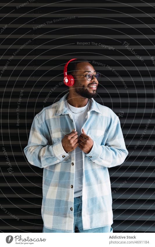 Lächelnder schwarzer Mann mit Kopfhörern und Jacke meloman Musik heiter Straße Gesang Großstadt Wand stylisch Afroamerikaner Glück männlich Typ Stil trendy