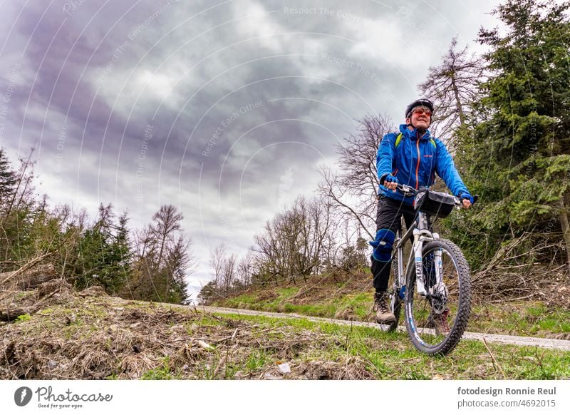 Mountainbiker am Rande eines geschotterten Waldwegs vor bewölktem Himmel. Moutainbike Fahrrad Biker Bäume Schotterweg Wolken