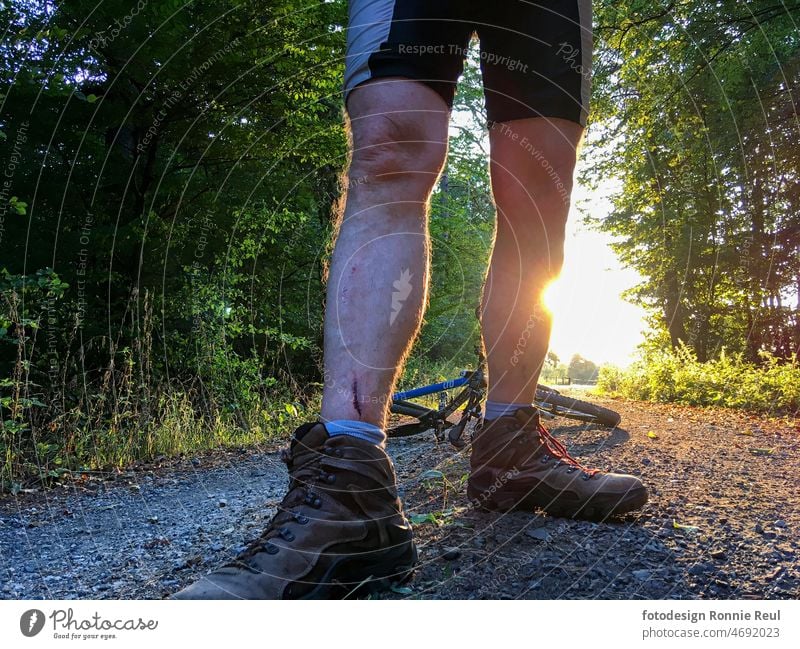 Verwundeter Mountainbiker im Gegenlicht auf einem geschotterten Waldweg. Fahrrad Unfall Sturz Wunde Beine Risiko Schmerz Schotter Bäume Sonne