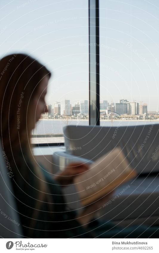 Frau liest ein Buch am Fenster sitzend mit Blick auf eine Stadtlandschaft in es an einem sonnigen Tag. Moderne casual lifestyle - unscharfe Silhouette der Frau mit einem Buch und Stadtlandschaft Blick in den Mittelpunkt. Lesen und Bildung.