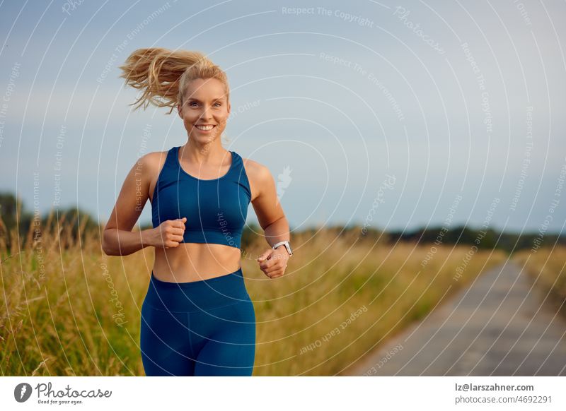 Fitte, gesunde junge Frau, die auf einer Landstraße joggt und mit einem glücklichen Lächeln voller Vitalität in einem aktiven Lebensstilkonzept an der Kamera vorbeigeht