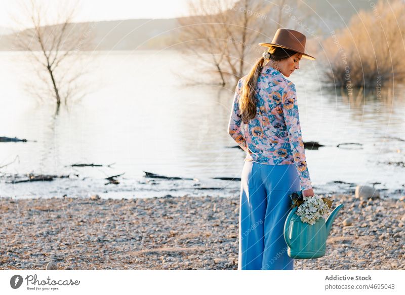 Frau mit Gießkanne mit Blumen am Ufer Strand Stil Wasser Fluss Natur feminin Flussufer Vorschein Hut Blütezeit Sommer Frühling Landschaft Küste Kopfbedeckung