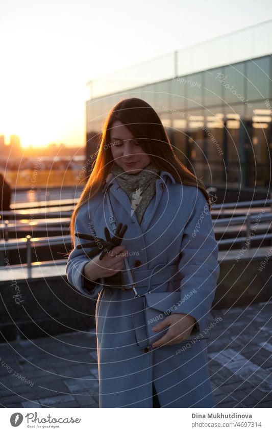 Frau in stilvollen blauen Mantel verlassen Büro bei Sonnenuntergang. Schöne attraktive junge Frau zu Fuß auf der Straße bei Sonnenuntergang oder Sonnenaufgang früh am Morgen in der Nähe der modernen Architektur Gebäude in der Stadt. Casual candid Lebensstil.