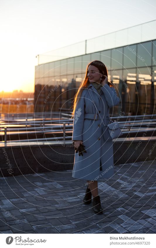 Frau in stilvollen blauen Mantel verlassen Büro bei Sonnenuntergang. Schöne attraktive junge Frau zu Fuß auf der Straße bei Sonnenuntergang oder Sonnenaufgang früh am Morgen in der Nähe der modernen Architektur Gebäude in der Stadt. Lässiger offener Lebensstil. Ganzkörperporträt.