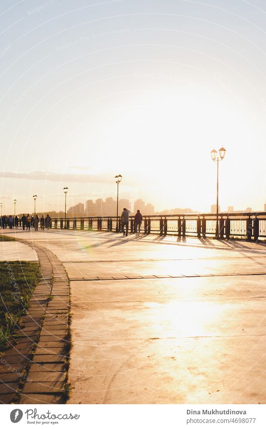 Stadtpromenade am Fluss bei Sonnenuntergang mit goldenem Licht gefüllt. Entfernte Silhouetten von Menschen, die auf dem Damm spazieren gehen. Sonnenaufgang Straßenansicht, Sonnenlicht reflektiert von der Straße. Städtische Szene hell und luftig Foto.