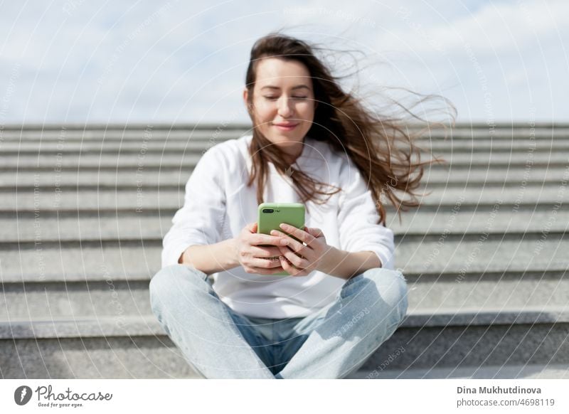 Junge Millennial-Frau in weißem Hemd, die ein Selfie macht oder mit ihrem Handy auf den Stufen eines Gebäudes videochattet. Weibliche Studentin, die auf den Treppenstufen eines Universitätsgeländes sitzt und Technologie nutzt. Echte Menschen mit Handys.