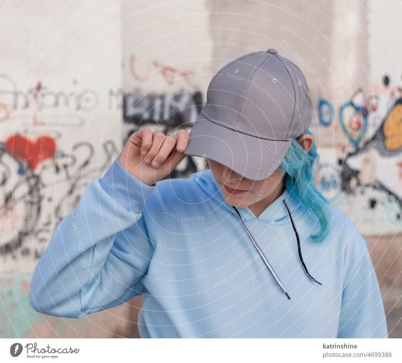 Blauhaariges Teenager-Mädchen in blauem Kapuzenpulli und Baseballmütze, das an einer Graffiti-Wand steht hell-blau Attrappe Verschlussdeckel blauhaarig