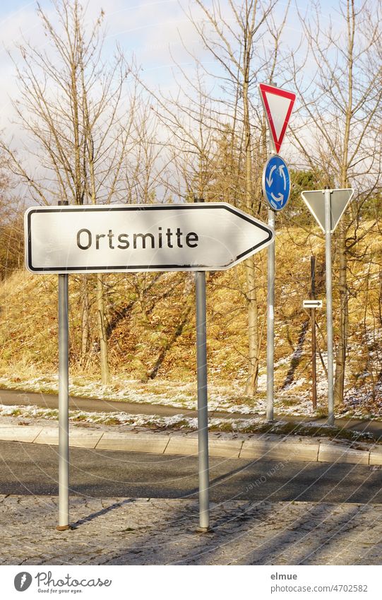 im Vordergrund auf einer Verkehrsinsel, nach rechts weisend, das Schild  - Ortsmitte - und im Hintergrund die Verkehrsschilder  - Kreisverkehr - und - Vorfahrt gewähren - bei frostigem Sonnenwetter