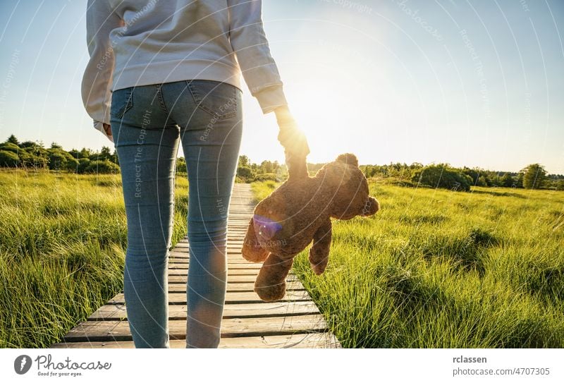 Frau steht auf einer Promenade und hält ein Teddybär-Spielzeug bei Sonnenuntergang, Platz für Ihren individuellen Text. Bär Rücken Erwachsener allein Weg