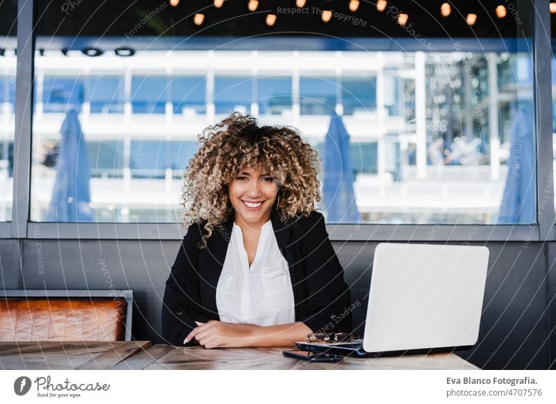 Lächelnde hispanische afroamerikanische Geschäftsfrau in einem Café, die an einem Laptop und einem Mobiltelefon arbeitet. Computer Afro-Look Frau Business