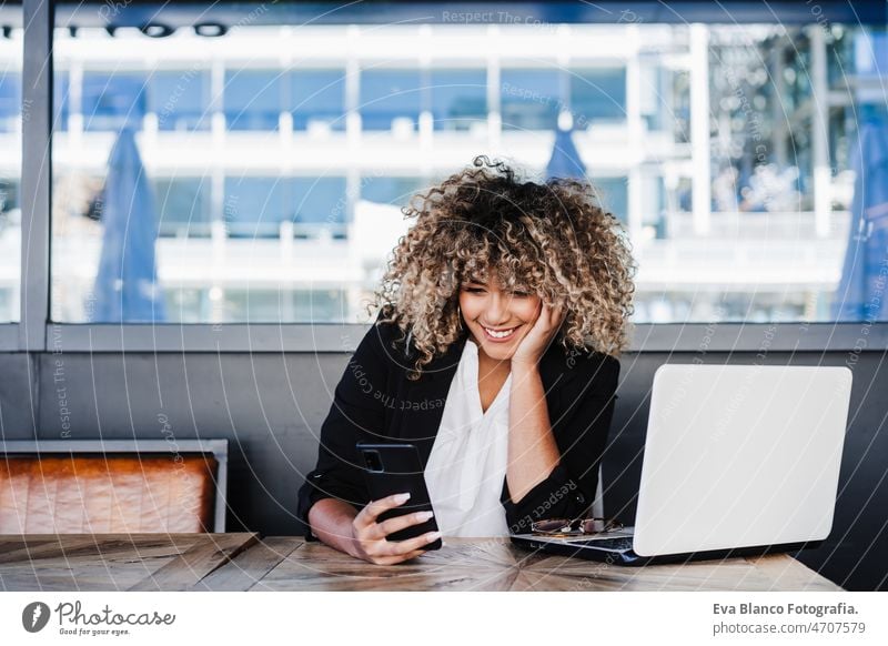 Lächelnde hispanische afroamerikanische Geschäftsfrau in einem Café, die an einem Laptop und einem Mobiltelefon arbeitet. Computer Afro-Look Frau Business