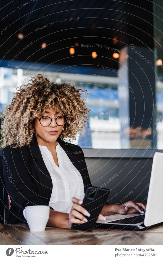 Lächelnde hispanische afroamerikanische Geschäftsfrau in einem Café, die an einem Laptop und einem Mobiltelefon arbeitet. Computer Afro-Look Frau Business