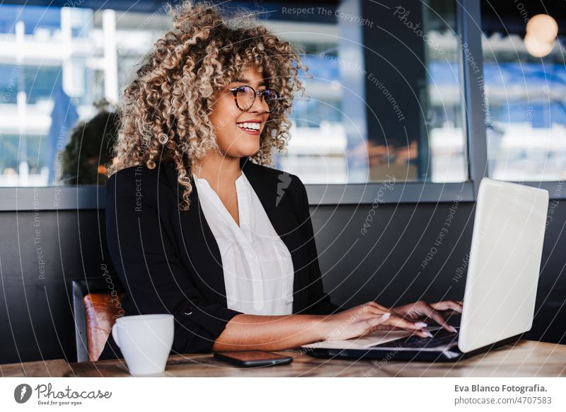 Lächelnde hispanische afroamerikanische Geschäftsfrau in einem Café, die an einem Laptop und einem Mobiltelefon arbeitet. Computer Afro-Look Frau Business