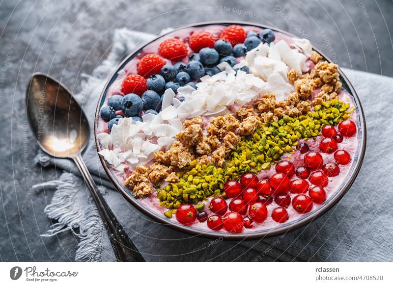 Gesunde Frühstücksbowl, mit Heidelbeer-Smoothie, Himbeeren, Heidelbeeren, Kokosnuss, Nüssen und Johannisbeeren als Topping auf einem Tisch mit Löffel