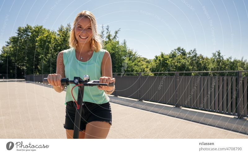 Junge Frau lächelnd bereit, einen Elektroroller vor einer Stadtlandschaft zu fahren, Platz für Ihren individuellen Text. Tretroller Großstadt Kick E-Roller