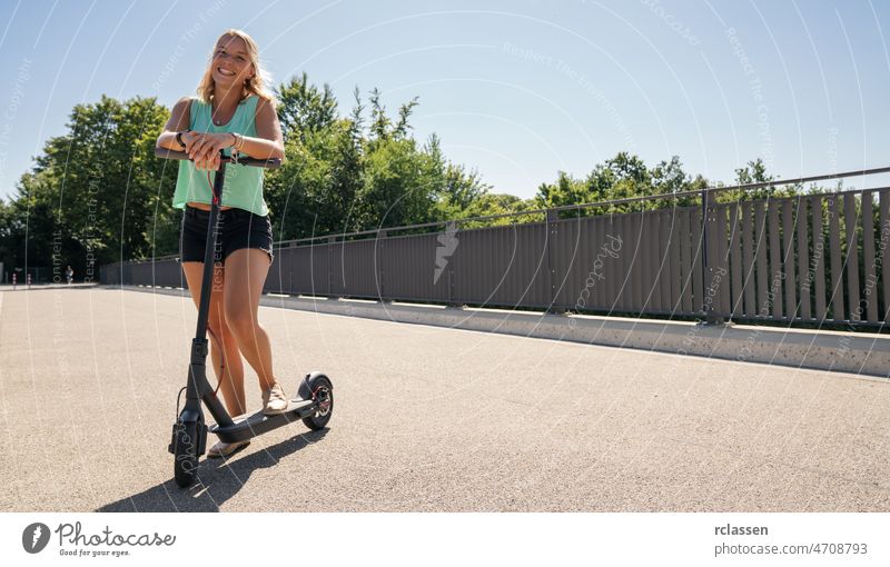 Junge Frau fährt auf einem schwarzen Elektroroller vor einer Stadtlandschaft, Platz für Ihren individuellen Text. Tretroller Großstadt Kick E-Roller