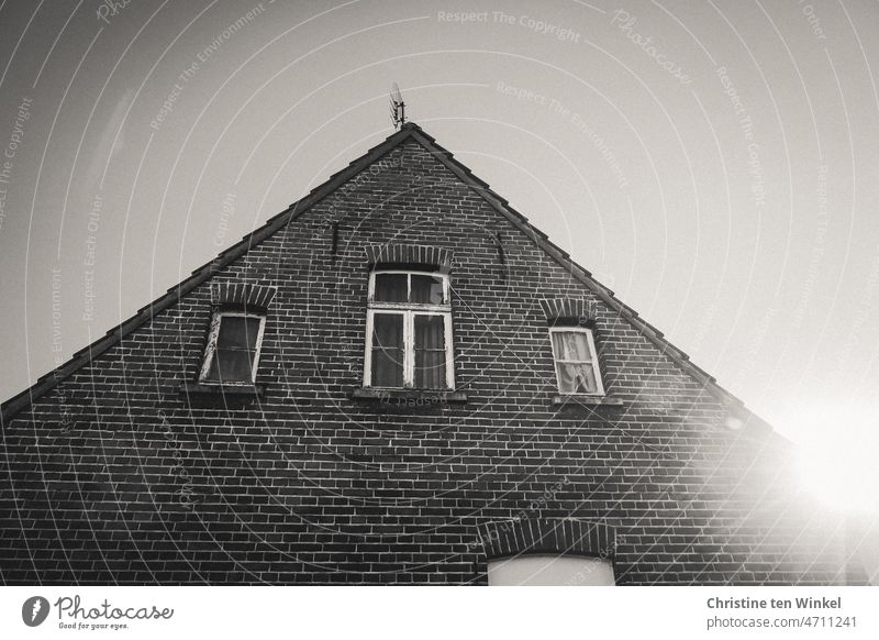 Düsteres unbewohntes altes Backsteinhaus. Drei Fenster und eine Antenne. Die Sonne kommt gerade hinter dem Dach hervor. Haus Gebäude Fassade Backsteinfassade