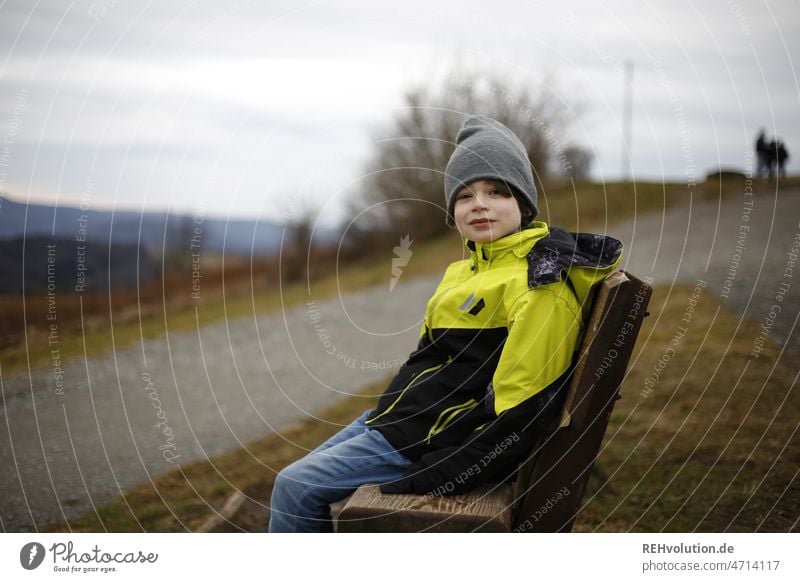 Kind sitzt auf einer Bank Mensch Junge Kindheit Außenaufnahme Jacke Landschaft natürlich authentisch Mütze Wetter Berge Natur windig Wolken 3-8 Jahre Wind