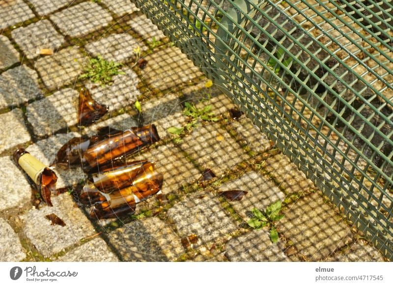 vor einer grünen Sitzbank aus Metall liegt auf den Pflastersteinen eine zerbrochene braune Glasflasche / Umweltverschmutzung / Glasscherben / Verletzungsgefahr