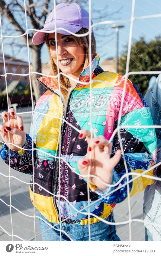 Frau, die sich am Zaun festhält und eine bunte Jacke trägt umklammert mehrfarbig Basketballplatz Verschlussdeckel Person im Freien schön Erwachsener attraktiv