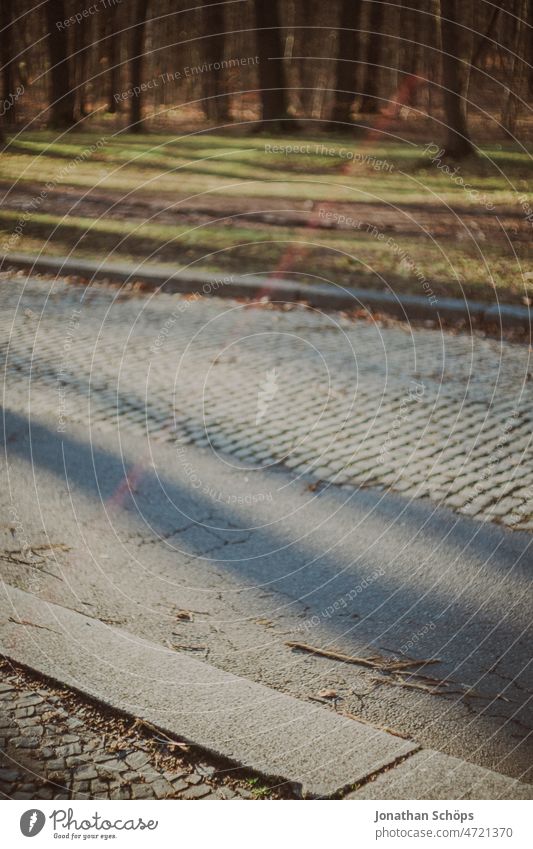 Bordsteinkante an einer leeren Straße am Wald Gehweg Fußweg Kopfsteinpflaster Flickenteppich Waldrand Sunflair Wiese grau Asphalt urban Straßenbelag Straßenrand