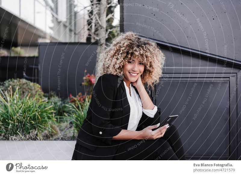 schöne lächelnde Business-Frau mit Handy sitzen auf Treppen in der Stadt. Gebäude Hintergrund Afro-Look hispanisch Geschäftsfrau Großstadt Wolkenkratzer jung