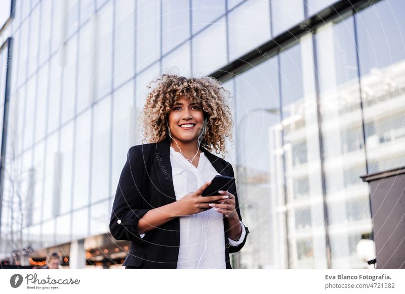 schöne lächelnde Geschäftsfrau mit Mobiltelefon und Kopfhörer in der Stadt. Gebäude Hintergrund Afro-Look hispanisch Handy Großstadt Wolkenkratzer Business jung