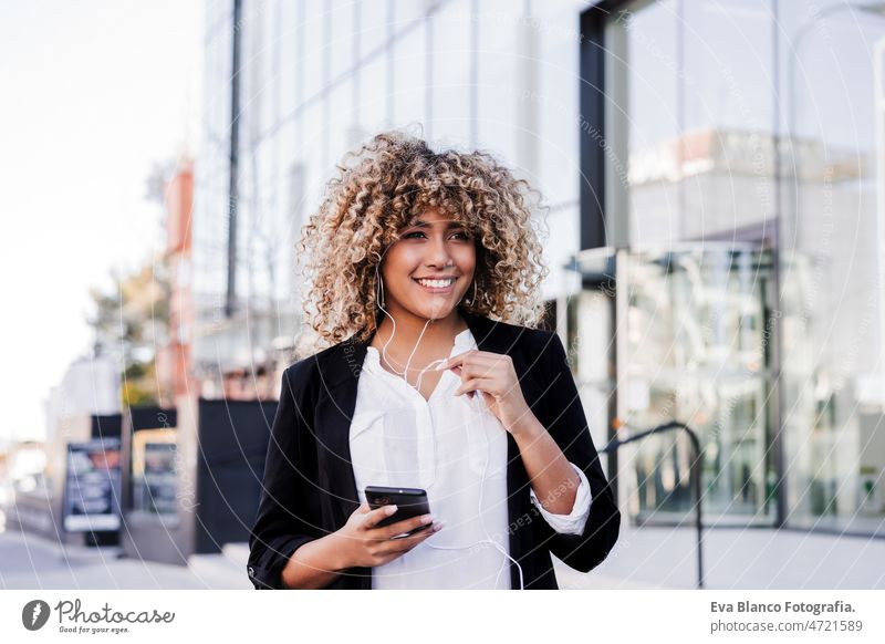 schöne lächelnde Geschäftsfrau mit Mobiltelefon und Kopfhörer in der Stadt. Gebäude Hintergrund Afro-Look hispanisch Handy Großstadt Wolkenkratzer Business jung