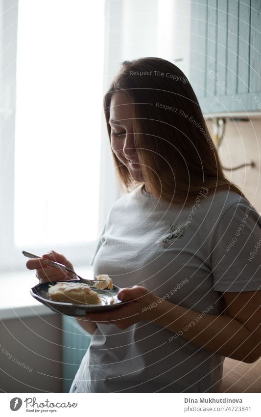 Junge Frau zu Hause, die morgens in der Küche stehend ein Dessert isst und in den neuen Tag hineinlächelt Morgen gute Miene zum bösen Spiel offen echte Menschen