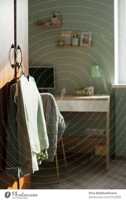 Gemütliches Zimmer einer jungen Frau mit auf dem Bügel hängenden Kleidern, Schreibtisch mit einem Personal Computer in grünen Pastellfarben. heimwärts Raum