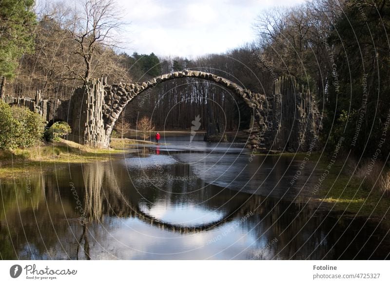 Unter der Rakotzbrücke steht Bellaluna mit ihrem roten Mäntelchen. Sie und die Brücke spiegeln sich auf der teils noch zugefrorenen Wasseroberfläche Park