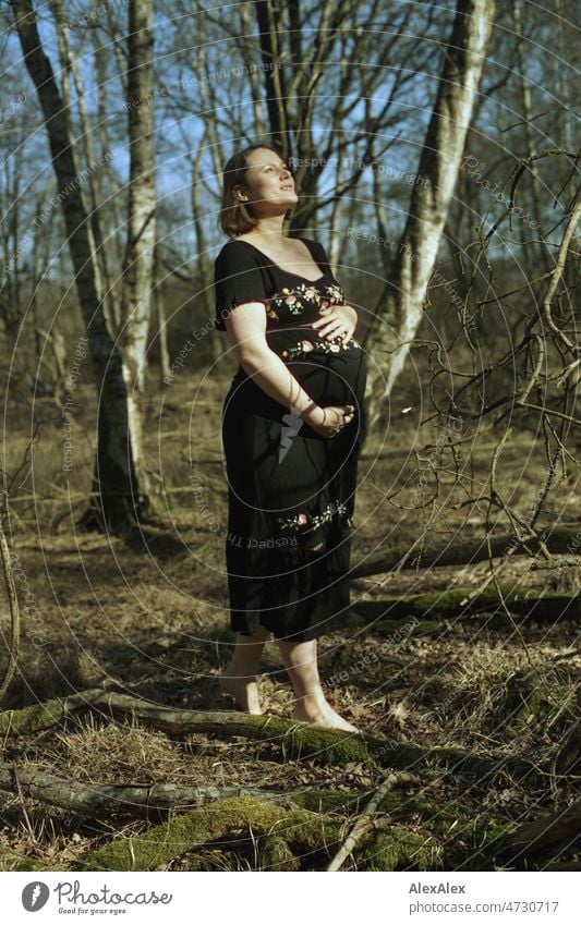 Analoges Ganzkörperportrait einer jungen, schwangeren Frau, die barfuß in einem Laubwald steht und sich den Babybauch mit einer Hand hält Lächeln kraftvoll Gras