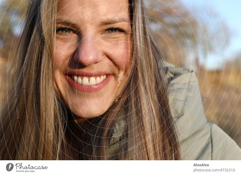 Große, blonde Frau mit langen Haaren in warmer, hellgrüner Jacke lächelt draussen sehr nah in die Kamera junge Frau schön langhaarig lange Haare Portrait nahe